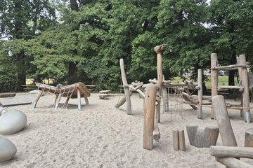 Holzspielplatz im Johannapark Leipzig mit Rutsche, Kletternetzen und Balancier-Elementen auf sandigem Untergrund, umgeben von Bäumen.