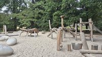 Holzspielplatz im Johannapark Leipzig mit Rutsche, Kletternetzen und Balancier-Elementen auf sandigem Untergrund, umgeben von Bäumen.