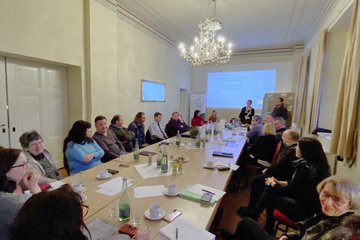 viele Menschen sitzen sin an einem großen Tisch und diskutieren, am Tischende ist einer Leinwand 