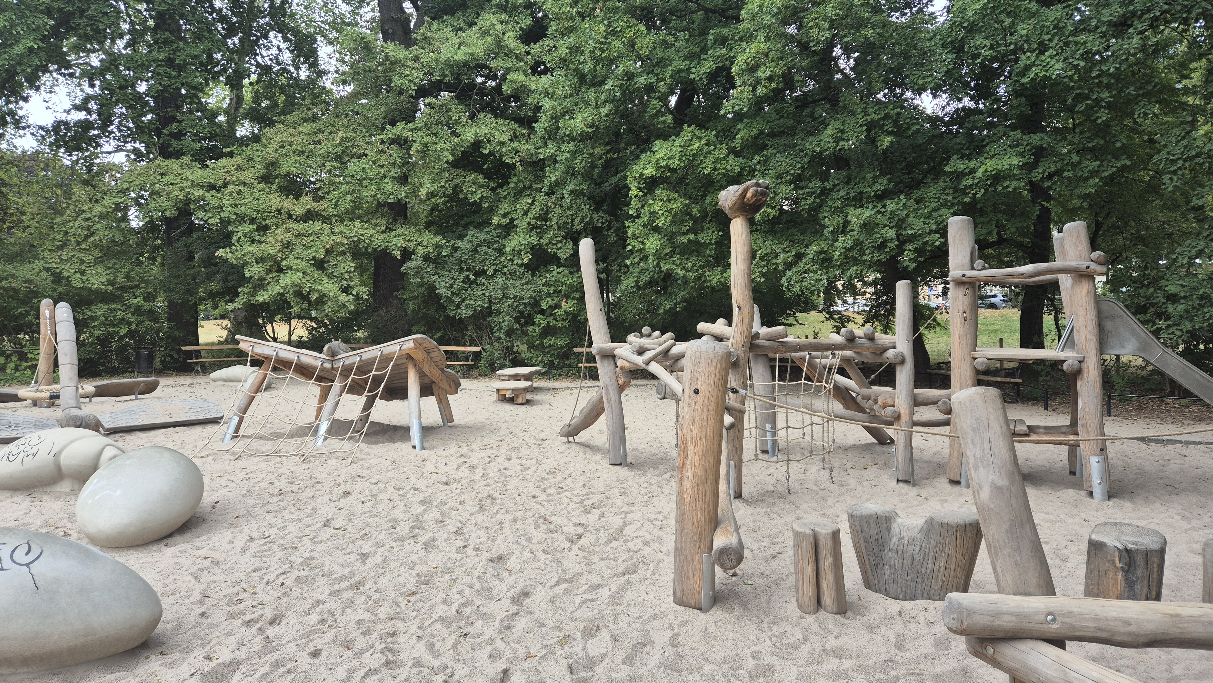 Holzspielplatz im Johannapark Leipzig mit Rutsche, Kletternetzen und Balancier-Elementen auf sandigem Untergrund, umgeben von Bäumen.