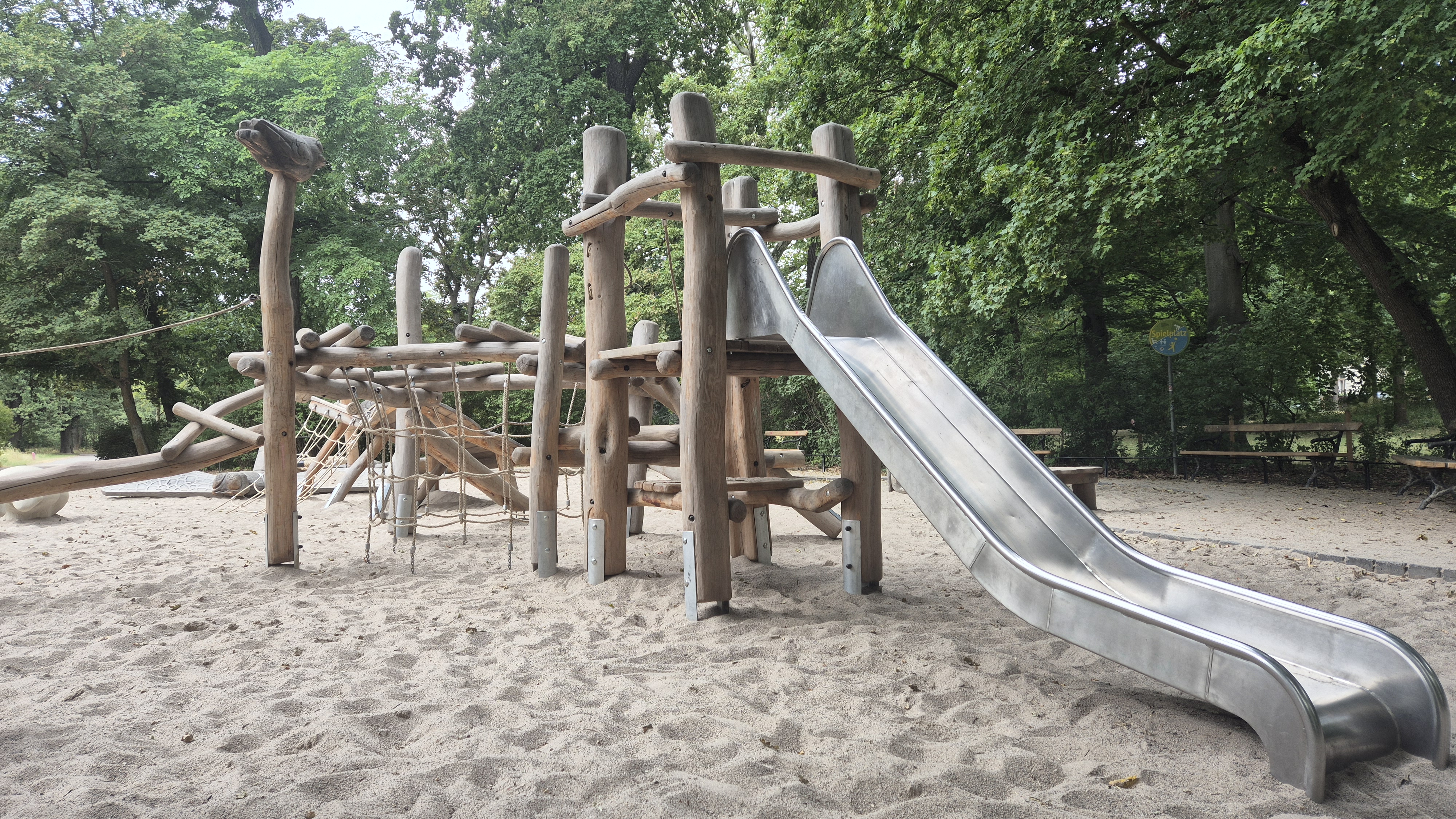 Große Edelstahlrutsche mit Holzkonstruktion auf einem Sandspielplatz im Johannapark, umgeben von Bäumen und Bänken.