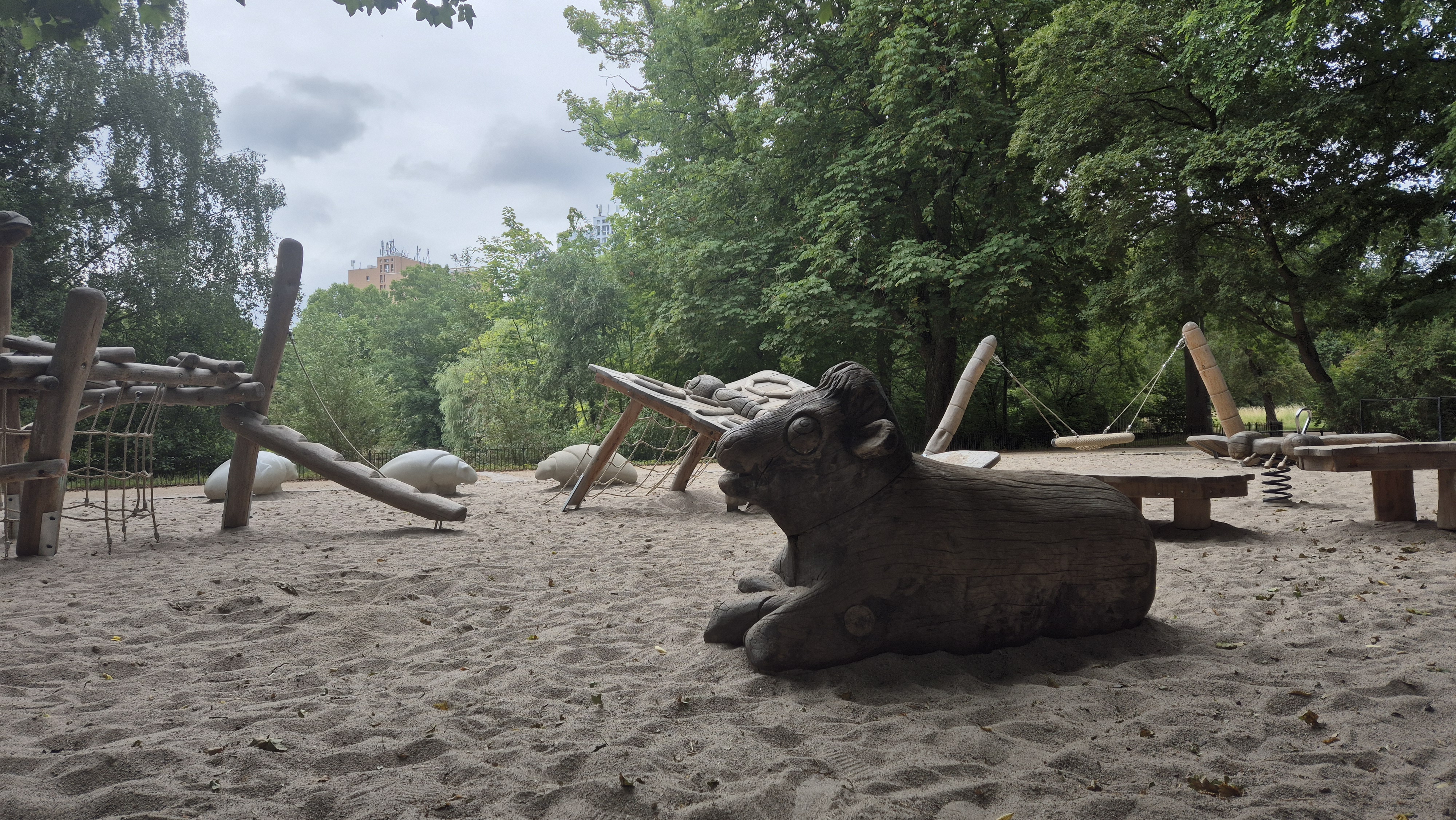 Spielplatz im Johannapark mit großen Holzfiguren in Tierform, darunter eine Kuh und eine Libelle, auf hellem Sandboden im Grünen.